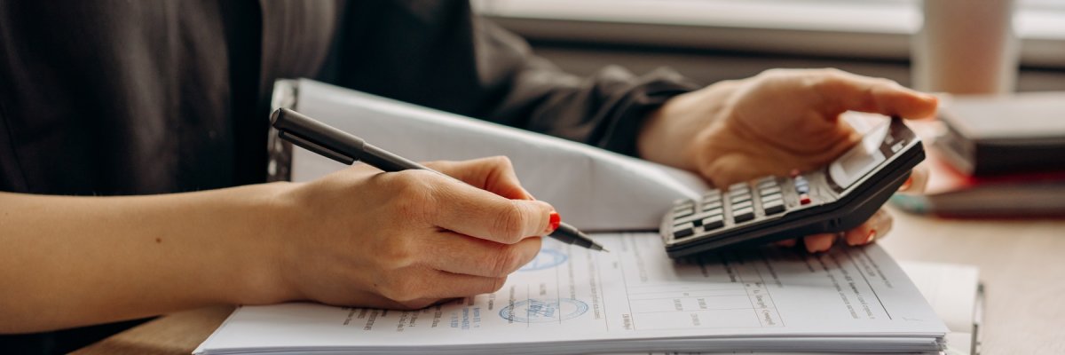 Ein offener Papierordner liegt vor einer Person, von der Person sind nur die Arme zu sehen, eine Hand hält einen Kugelschreiber die andere einen Taschenrechner