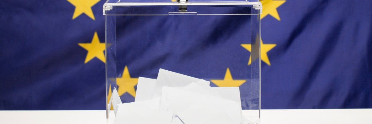 Transparente Wahlurne mit weißen Briefumschlägen vor einer Europaflagge