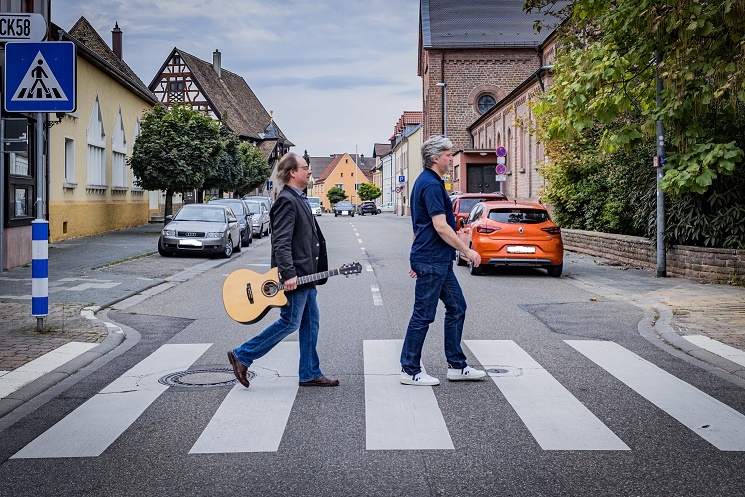 Die Musiker Alexis und Gereon (mit Gitarre in der rechten Hand) laufen über einen Zebrastreifen