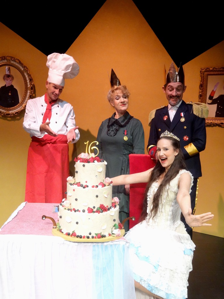 Dornröschen sitzt am Tisch, auf dem Tisch ist eine dreistöckige Geburtstagstorte zum 16. Geburtstag abgestellt. Im Hintergrund stehen die Eltern (das Königspaar) und der Tortenbäcker.