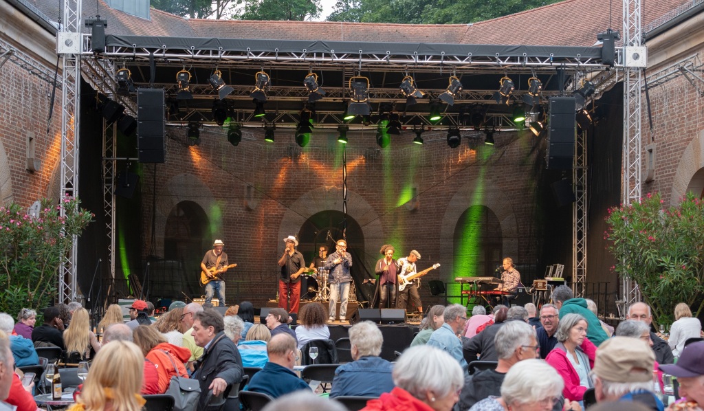 Die Band auf der Bühne im Hufeisen, vor der Bühne sind viele Bistrotische mit Besuchern besetzt.
