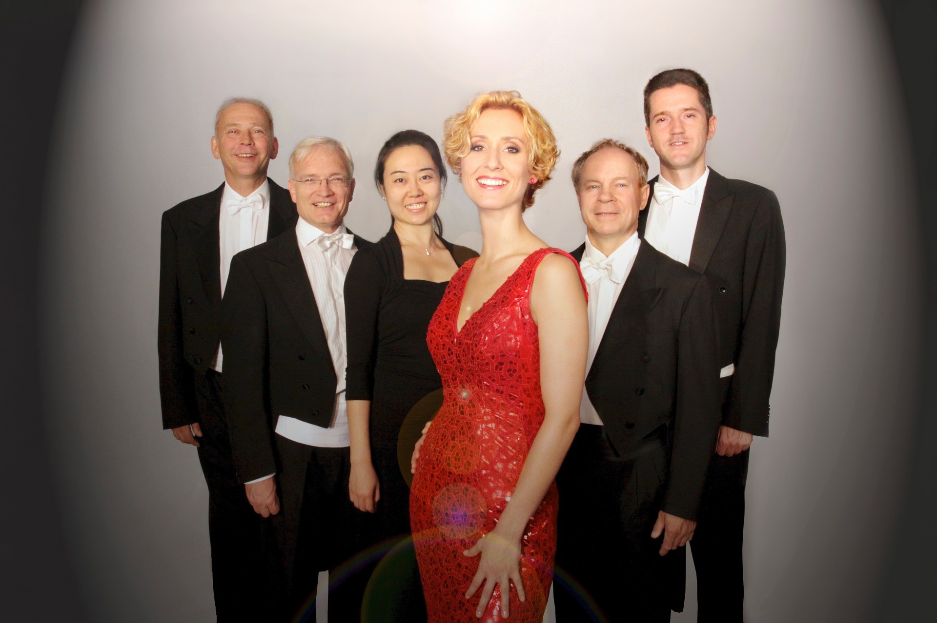 Bild des Ensemble "Musica Varia Ensemble", im Vordergrund steht die Sängerin im roten Kleid, dahinter stehen die Musikerinnen und Musiker (ohne Instrumente) in dunkler Kleidung.