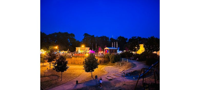 Blick auf das historische Festungsgebäude mit der Bühne des Vereins Interkultur, nachts mit bunten Lichtern beleuchtet, mit vielen Besuchern, im Vordergrund sind noch Teile des Spielplatz im Park "Fronte Lamotte" zu sehen