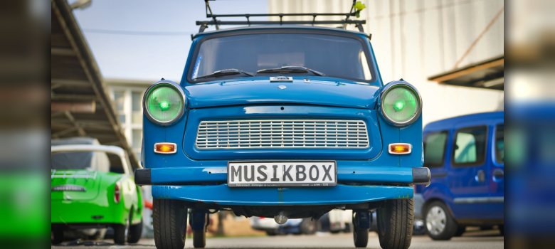 Frontansicht eines blauen Trabbi mit grünen Scheinwerfern. Auf dem Nummernschild steht der Text "Musikbox".
