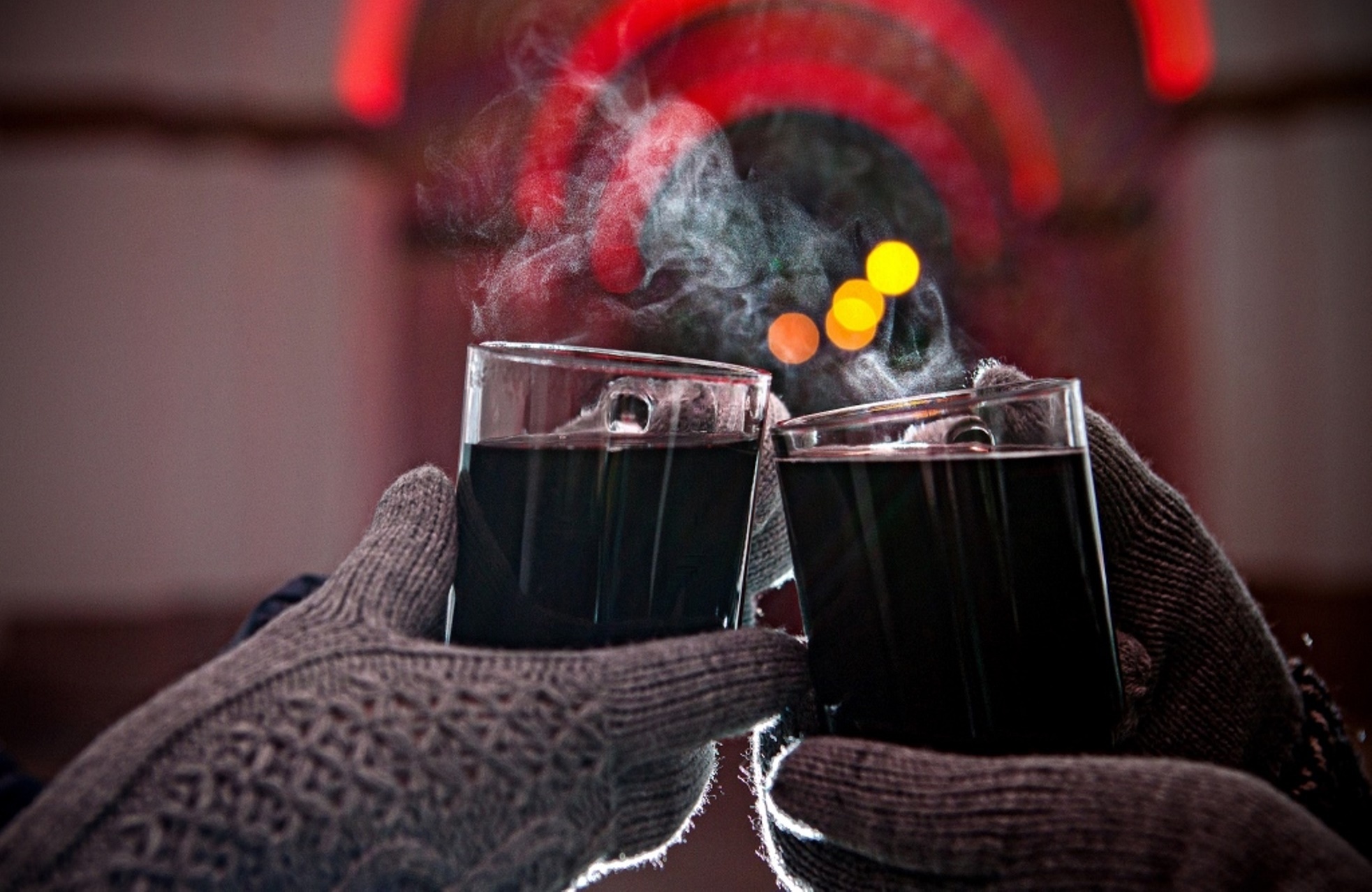 Zwei Tassen dampfender Glühwein stoßen an. Die Hände an den Gläsern haben graue Strickhandschuhe an. Im Hintergrund ist der beleuchtete Durchgang am Weißenburger Tor zu sehen.