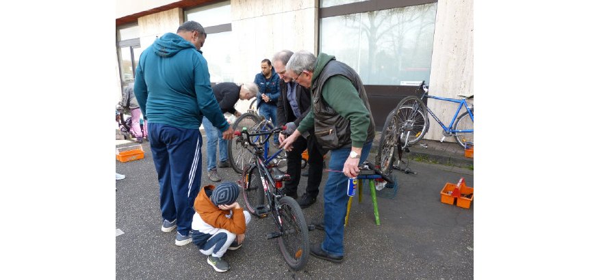 Ein Kinderfahrrad wird begutachtet, was alles repariert werden muss / kann. Mehrere Männer stehen um das Fahrrad herum, ein Junge sitzt daneben.