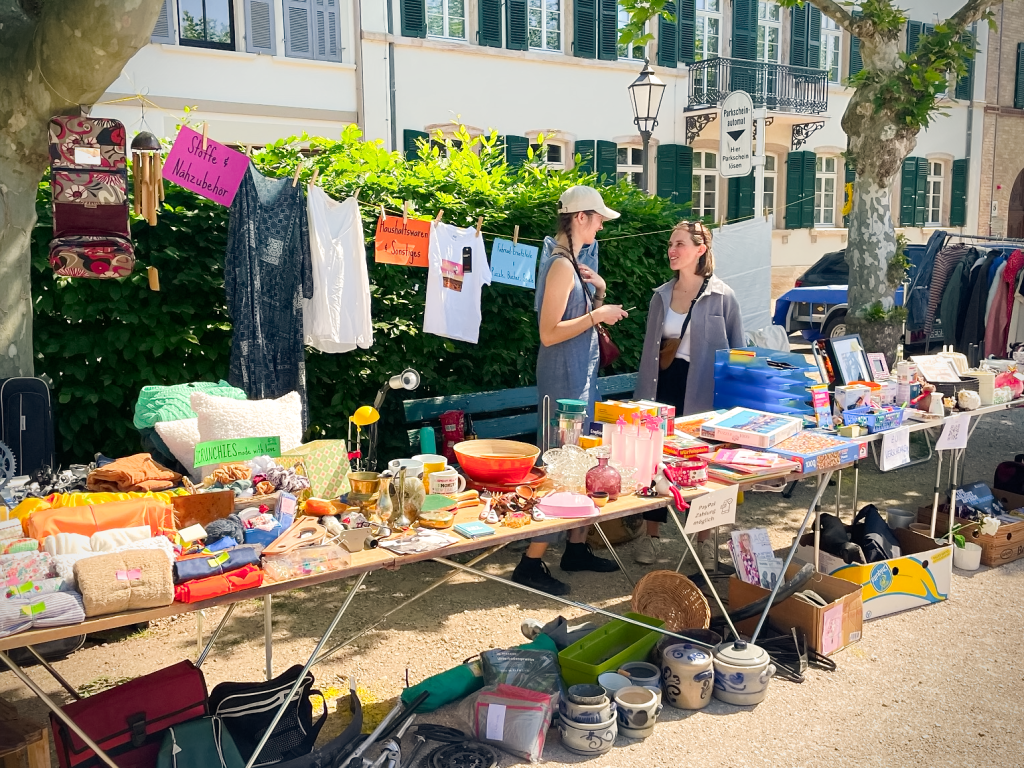 zwei Frauen hinter einem Flohmarkttisch mit ganz verschiedenen Angeboten. Im Hintergrund sind Hecken und Bäume zu sehen.