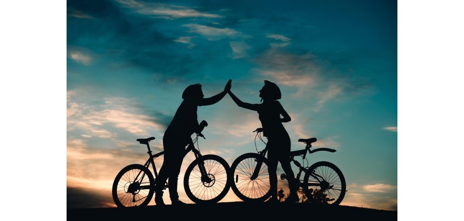 Zu sehen sind die Sillouetten zweier Fahrradfahrer, die sich gegenüber stehen und sich "abklatschen", vor einem Sonnenuntergang.
