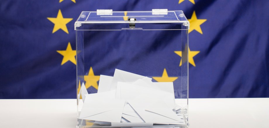 Transparente Wahlurne mit weißen Briefumschlägen vor einer Europaflagge